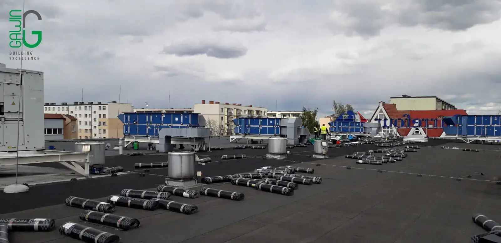 Płaski dach z rozłożonym zwijanym materiałem dachowym; w tle widoczne są budynki i zachmurzone niebo.