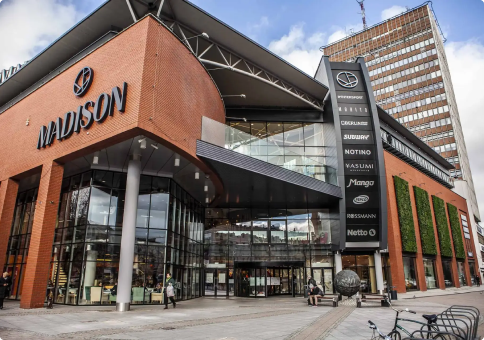 Nowoczesne centrum handlowe z dużym szyldem „MADISON”, szklanym wejściem, widocznymi logami sklepów i wysokim budynkiem biurowym w tle.