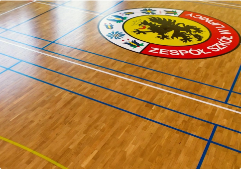 Podłoga sali gimnastycznej z różnymi liniami boisk sportowych i dużym okrągłym emblematem z napisem „ZESPÓŁ SZKÓŁ W LIPNICY” przedstawiającym czarnego gryfa i elementy dekoracyjne.