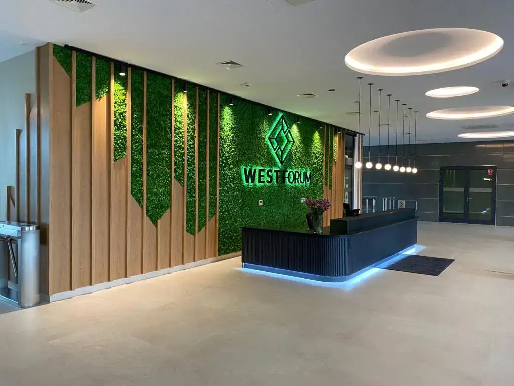 Nowoczesne lobby biurowe z zieloną ścianą, drewnianymi akcentami, czarnym biurkiem recepcyjnym z niebieskim oświetleniem, lampami wiszącymi i szyldem „West Forum”.